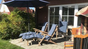 A patio with two lounge chairs and a table, a large umbrella, and a brick house with a tiled roof. - Ferienwohnung, Garding (Garding)