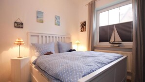 A bedroom with a bed, two bedside tables, a window with a sailboat decoration, and wall art. - Ferienwohnung, Garding (Garding)