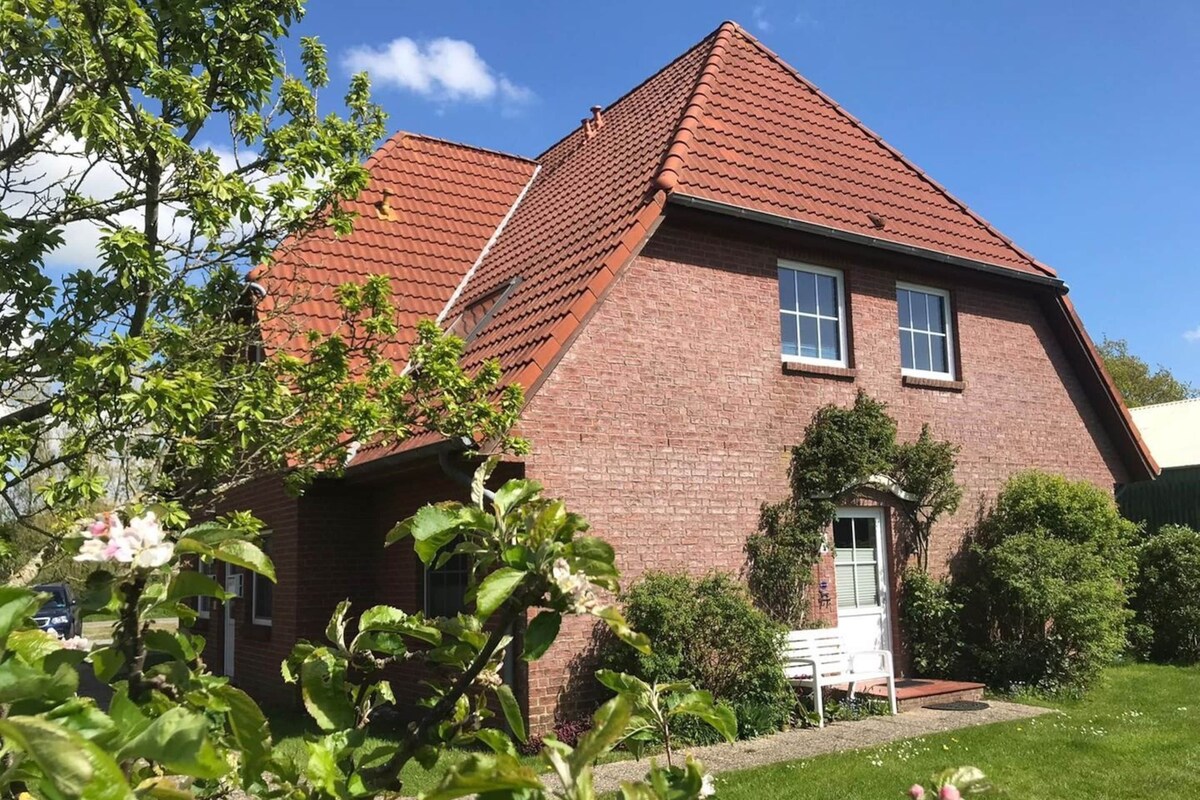 Ein zweistöckiges Backsteinhhaus mit rotem Ziegeldach, einer Haustür und einer kleinen Veranda mit einer Bank.