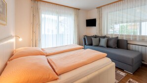 A hotel room with a bed, a sofa, a TV mounted on the wall, and large windows with sheer curtains. - Apartment in Fugen Near Spieljochbahn (Fuegen)