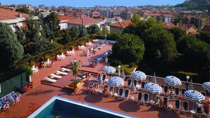 Zona de piscina en la azotea con sillones de descanso y sombrillas, rodeada de árboles y edificios.