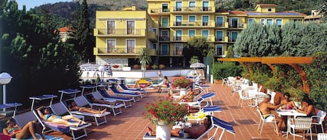 A rooftop pool area with sun loungers and tables, surrounded by buildings and trees.