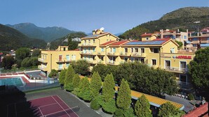 Un hotel de varios pisos con balcones, una cancha de tenis y un paisaje montañoso al fondo.