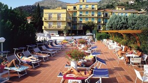 Un área de piscina en la azotea con tumbonas y mesas, rodeada de edificios y árboles.