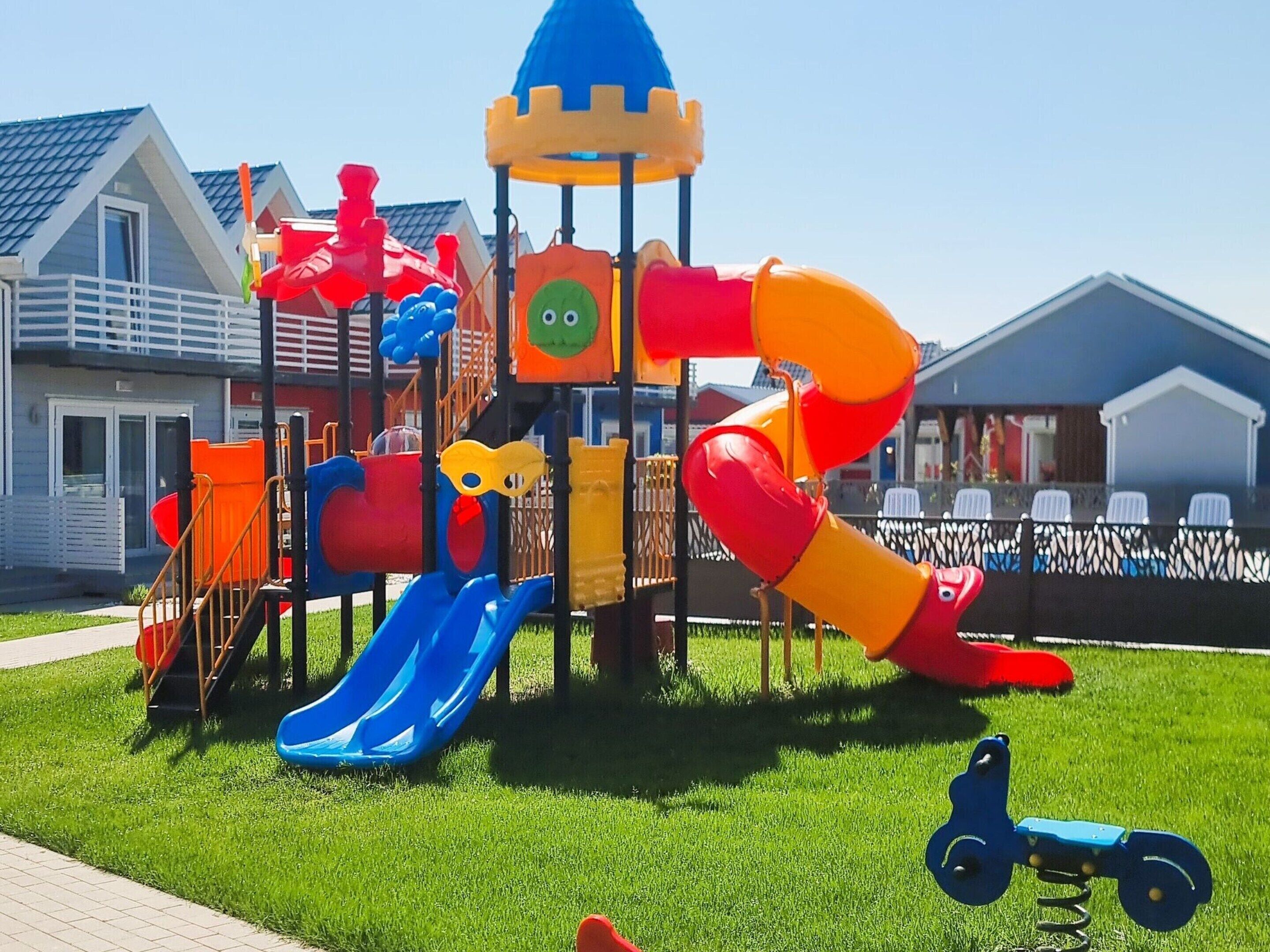 A playground with slides and climbing structures, surrounded by a fenced area and residential buildings in the background.