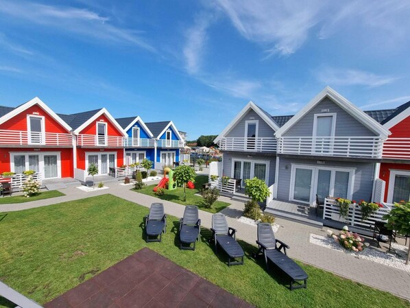 A row of colorful vacation cottages with a grassy lawn and lounge chairs in the foreground. - Two-story Houses With Terraces, Pool, Playground (Mielno)