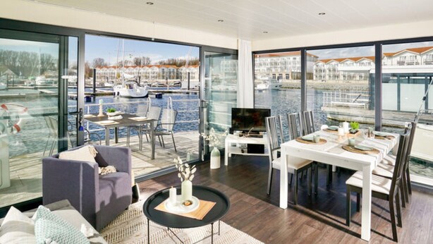A modern living room with a large glass wall, a dining area, and a view of a marina with boats. - Hausboot in Boltenhagen (Boltenhagen)