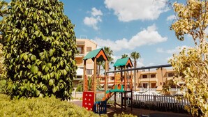 A playground with a swing set and climbing structures, surrounded by well-maintained bushes and palm trees, with a building in the background. - Mazarron Coastal Apartment for 3 (Mazarrón)