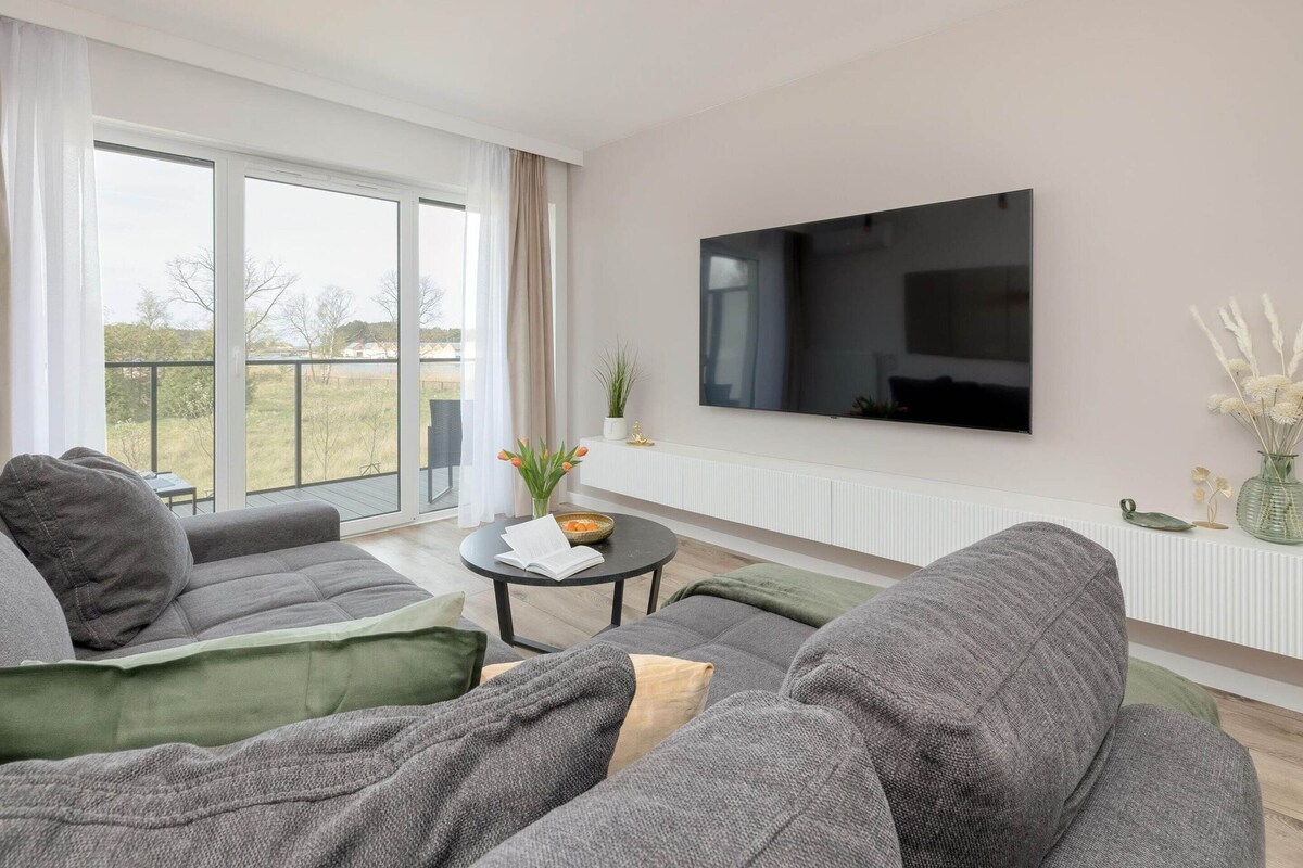 Un salon moderne avec des canapés gris, un téléviseur à écran plat et une grande fenêtre donnant sur l’extérieur.