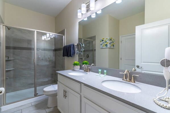 A modern bathroom with a glass-enclosed shower, two sinks, a large mirror, and a door leading to another room. - Spacious Luxury Champions Gate Retreat w/ Pool, Spa & Game Room – Sleeps 20 (Davenport)