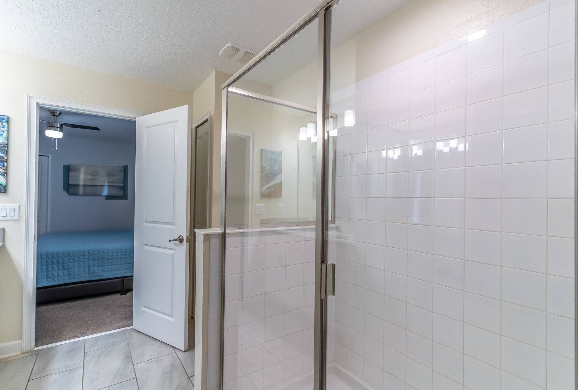 Une salle de bain moderne avec des carreaux blancs, une douche en verre et un lit visible dans la pièce adjacente.