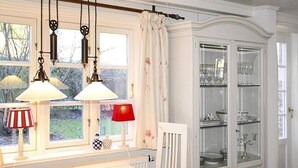 A kitchen with a white countertop, hanging pendant lights, and a red lamp. There is a white chair and a glass cabinet with dishes. - Ferienwohnung in Oevenum auf Foehr (Oevenum)