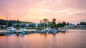 Marina con varias embarcaciones amarradas, un moderno complejo de edificios al fondo y una masa de agua tranquila que refleja el cielo.