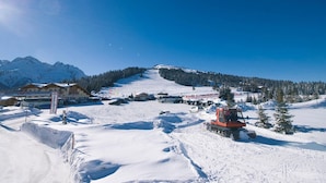 A snow-covered ski resort with a mountain backdrop, a red snow grooming machine, and a building with a sign.