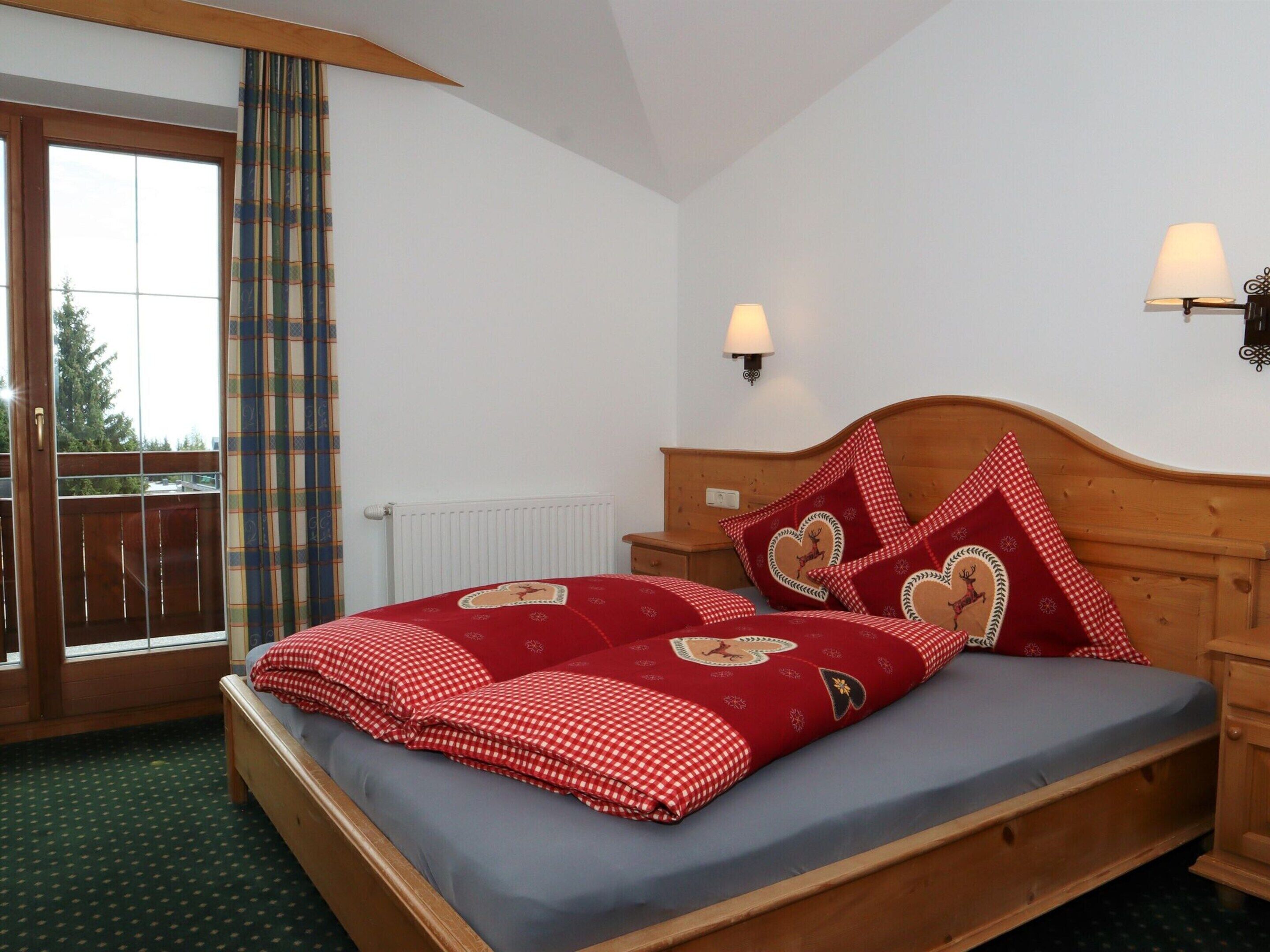 Une chambre avec un lit en bois, une literie à carreaux rouges, des oreillers en forme de cœur, une fenêtre donnant sur un beau paysage et une lampe fixée au mur.