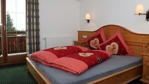 A bedroom with a wooden bed, red checkered bedding, heart-shaped pillows, a window with a view, and a wall-mounted lamp.