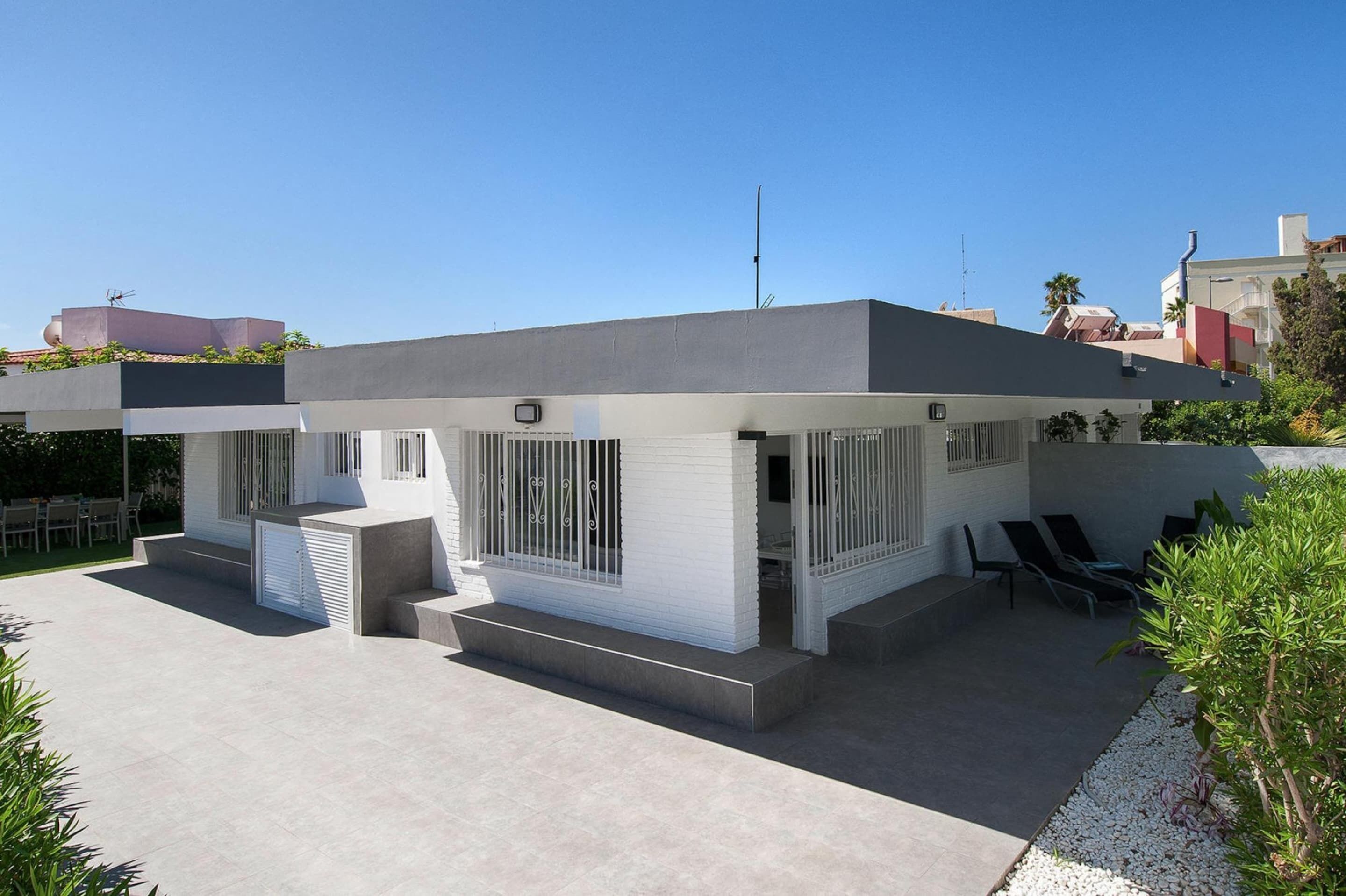 Une maison moderne avec un toit plat, de grandes fenêtres et un patio aménagé avec du mobilier d’extérieur.