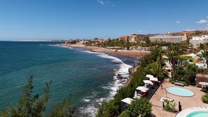 Un hotel frente al mar con vista a la playa, piscina y paseo marítimo con sombrillas.