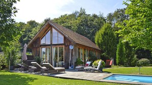 A wooden cabin with a large glass window, a swimming pool, and outdoor seating. - Chalet in Styria With Pool & Panoramic Views (Fehring)