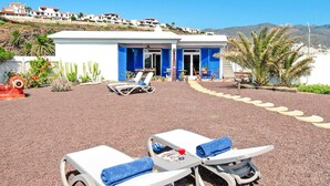 Un hôtel situé en bord de mer, avec une façade blanche et bleue, de nombreux fauteuils de plage et une vue sur la mer et les collines en arrière-plan.