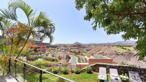 A balcony with a view of a landscaped garden, a pool area with lounge chairs, and a distant view of a hilly landscape. - Holiday Home, Maspalomas (San Bartolomé de Tirajana)