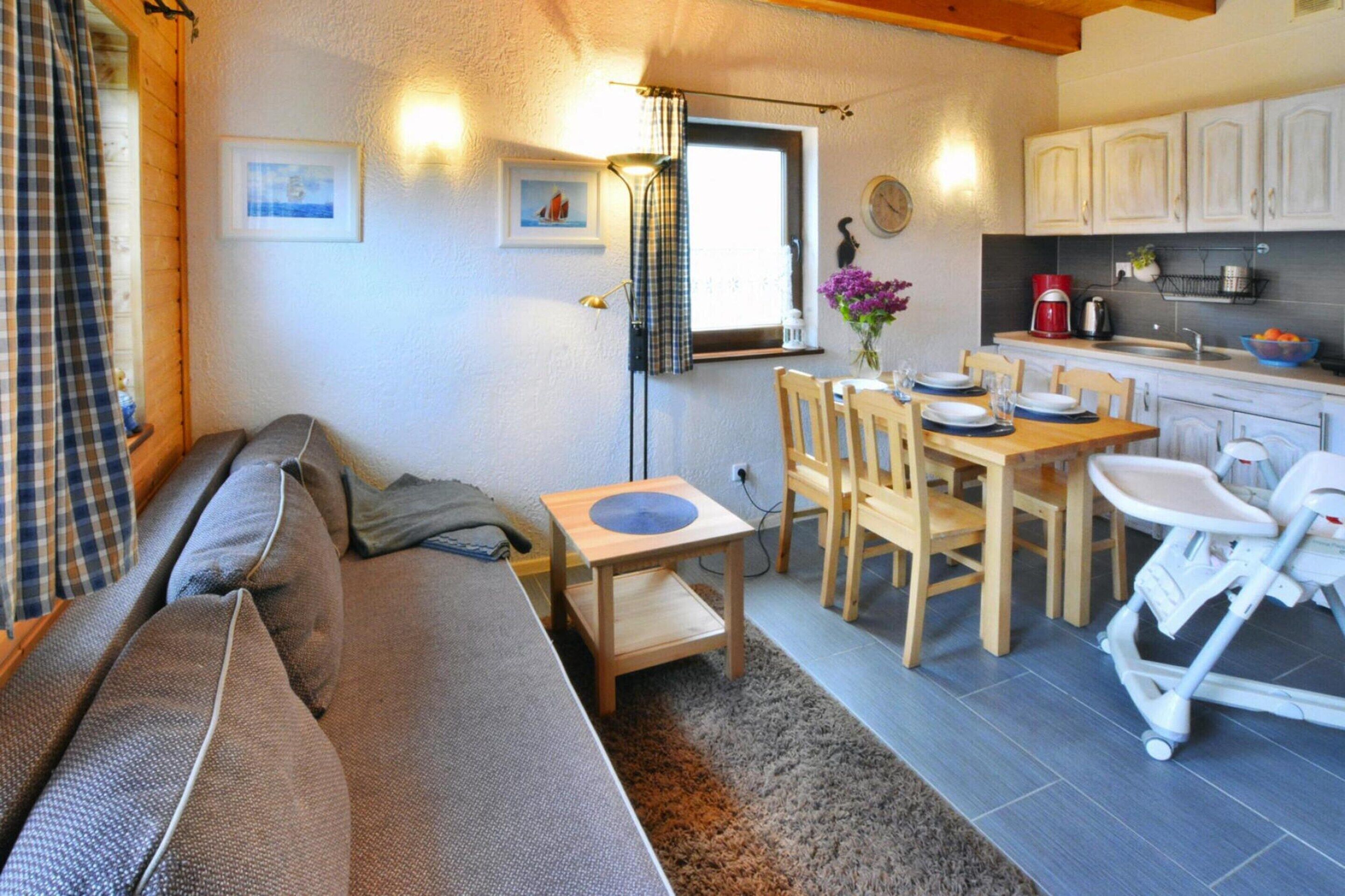 A cozy living room with a sofa, a small wooden table, and a dining area with chairs and a table. There is a window with curtains, a wall-mounted lamp, and a kitchen area with cabinets and a sink.