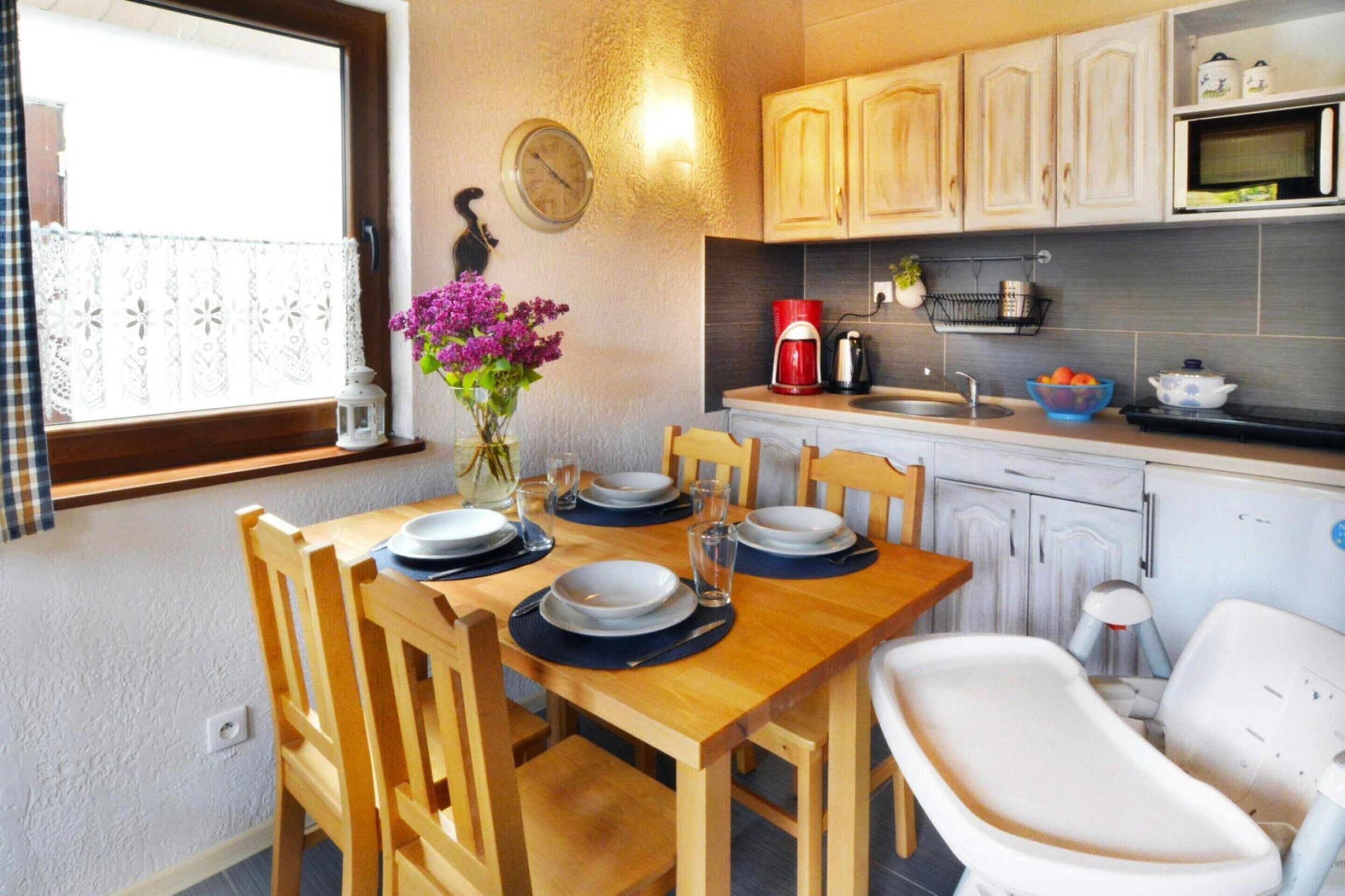 A kitchen with wooden cabinets, a dining table set for four, a clock on the wall, and a window with lace curtains.