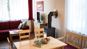 Un salon chaleureux avec un canapé rouge, des chaises en bois, une table à manger ornée d’un saladier de fruits et de verres, un téléviseur, une plante et un poêle à bois.