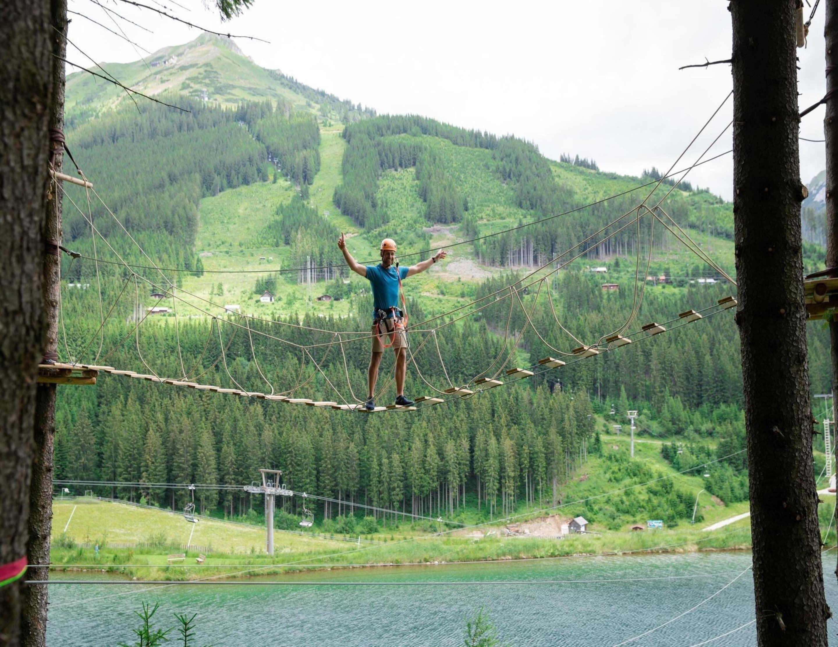 Une personne se trouve sur un pont de corde qui enjambe une étendue d’eau, avec un paysage montagneux en arrière-plan.