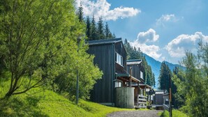 Une cabane en bois, accessible par un chemin de gravier, entourée d’une végétation luxuriante et avec des montagnes en toile de fond.