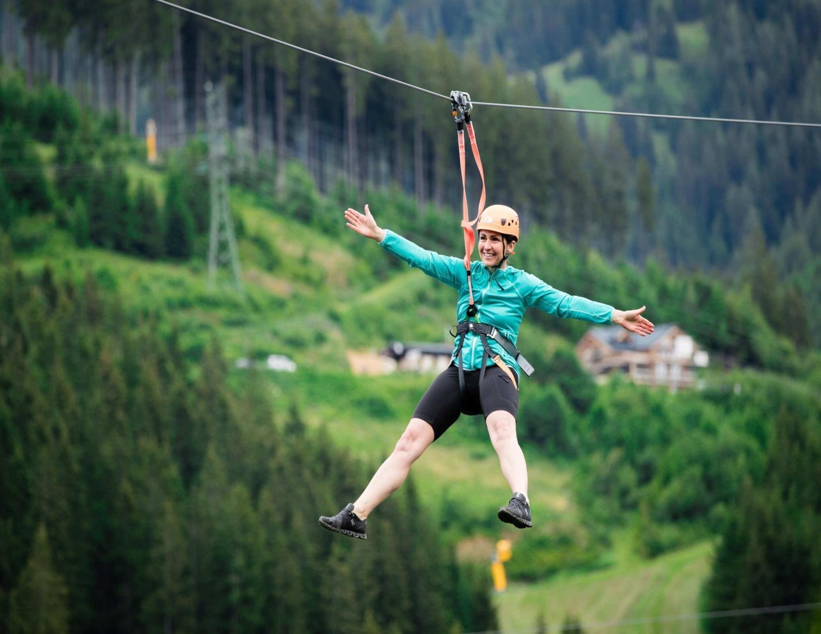 A person zip-lining with a harness and helmet, surrounded by a mountainous landscape with greenery and buildings in the background.