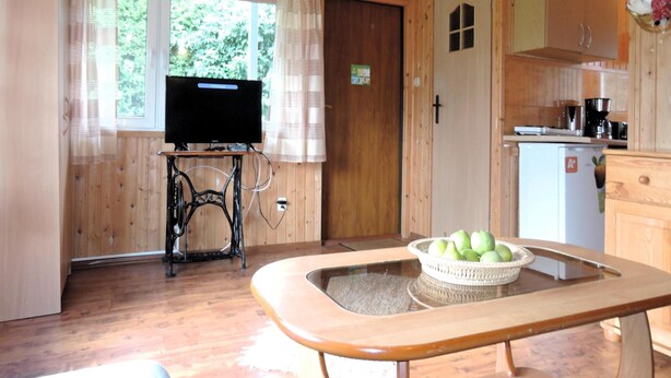 Interior de una cabaña de madera con una máquina de coser, un televisor, una zona de cocina y una mesa de comedor con una cesta de manzanas.