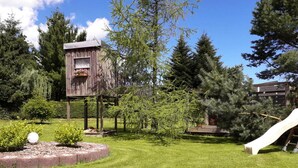 Una casita de madera con un macetero de flores, rodeada por un patio cubierto de césped, un tobogán y árboles.