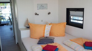 A bedroom with two beds, each with striped bedding and orange pillows. There is a small window with blinds, a door with a sign above it, and a chair visible in the background. - Hausboot in Boltenhagen (Boltenhagen)