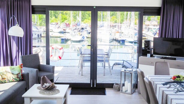 A modern room with a view of a marina, a sofa, a coffee table, a TV, and a dining area. - Hausboot in Boltenhagen (Boltenhagen)