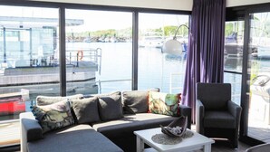 A modern living room with a sofa, armchair, and coffee table, large windows with a view of a marina and boats. - Hausboot in Boltenhagen (Boltenhagen)
