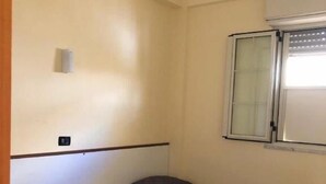 A small, single-room accommodation with two beds, a window with blinds, a wall-mounted air conditioning unit, and a small electrical outlet. - Baia dello Stretto (Reggio Calabria)