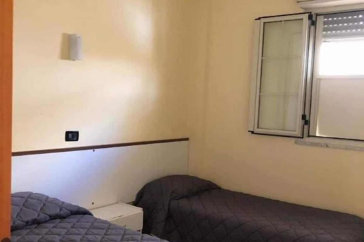 A small, single-room accommodation with two beds, a window with blinds, a wall-mounted air conditioning unit, and a small electrical outlet.