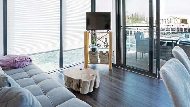 En moderne stue med en grå sofa, et træbord og et fjernsyn på et træstel. Store vinduer og en balkon med udendørs møbler er synlige.