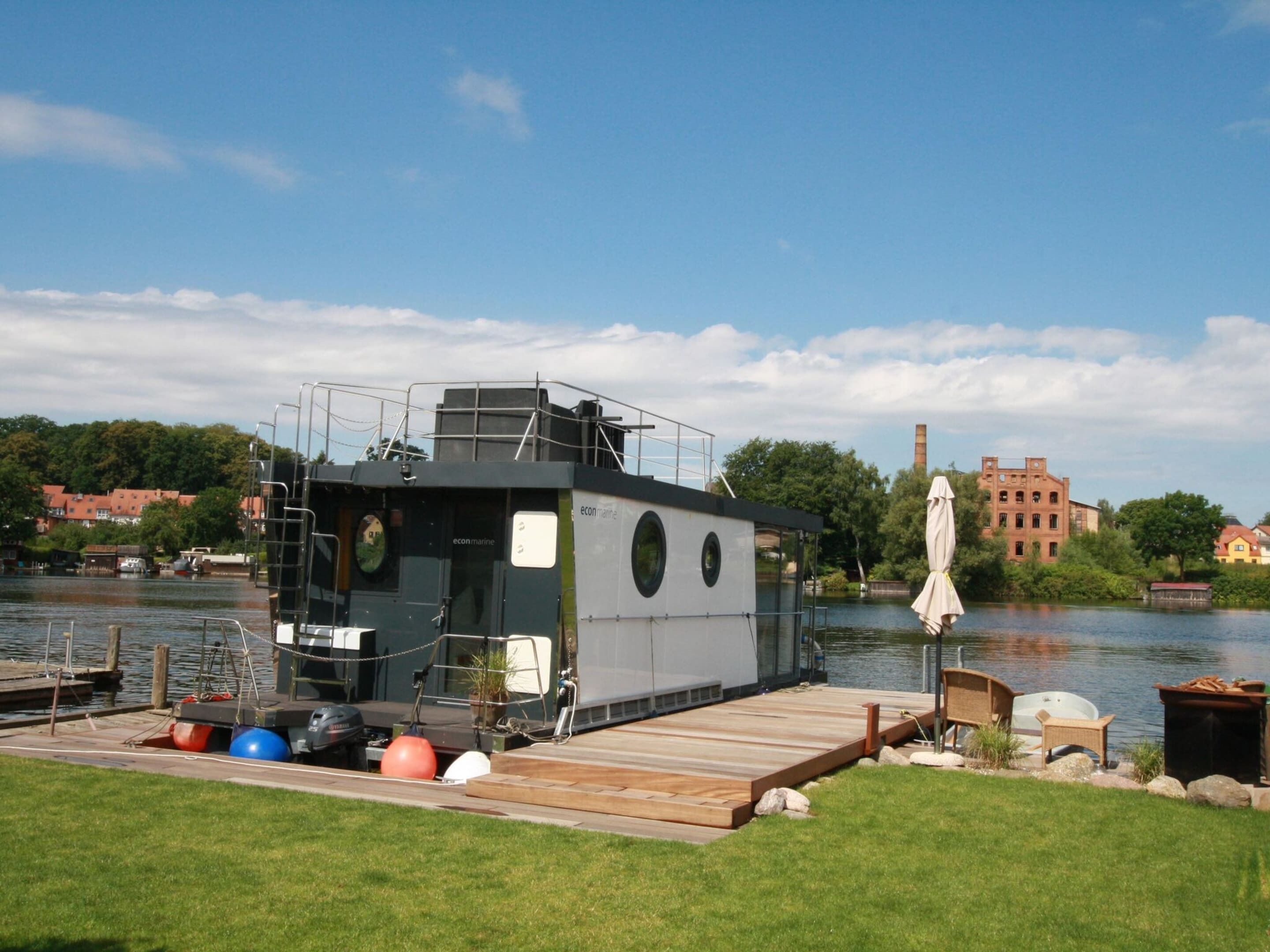 Ein Hausboot liegt an einem Steg, im Vordergrund befindet sich eine Rasenfläche und im Hintergrund sind Gebäude zu sehen.