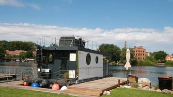 Ein Hausboot liegt an einem Steg, im Vordergrund befindet sich eine Rasenfläche und im Hintergrund sind Gebäude zu sehen.