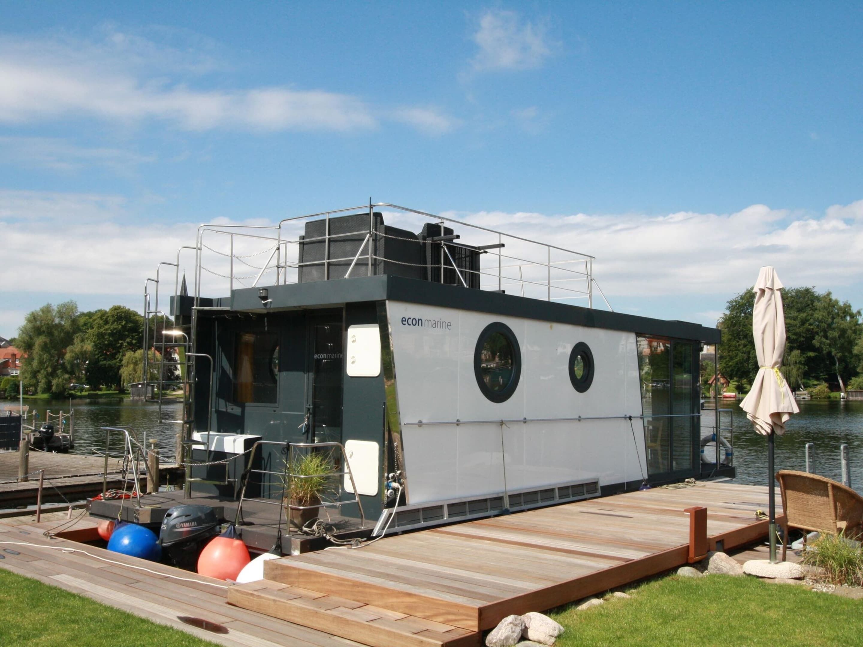 Ein Hausboot an einem Steg, mit einer Holzterrasse, einer kleinen Sitzgruppe im Freien und einem Sonnenschirm.
