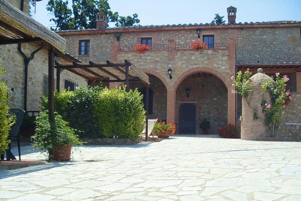 A courtyard with stone flooring, a pergola, and a building with arched doorways and flower boxes.