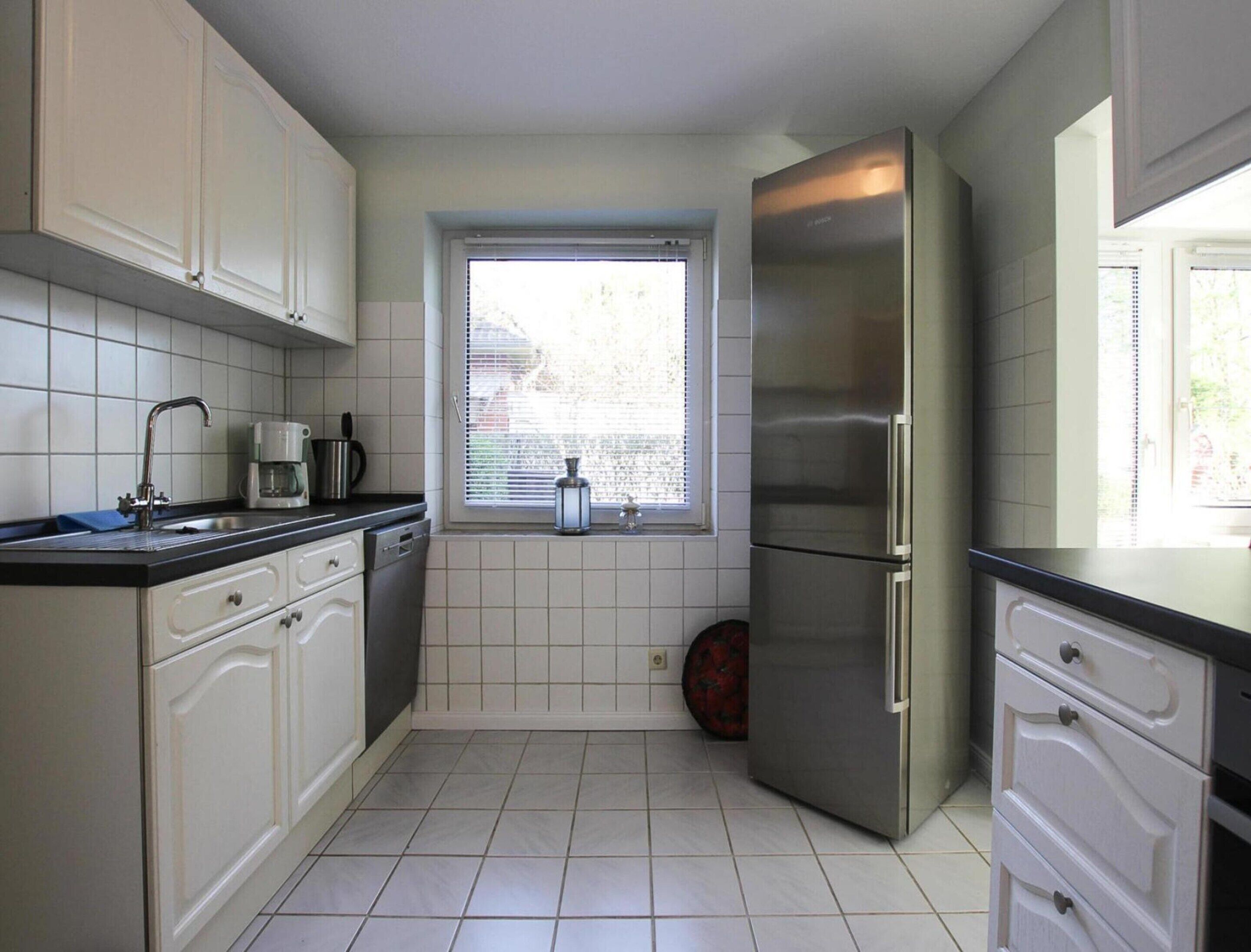 Una cocina pequeña con armarios blancos, un refrigerador de acero inoxidable, una ventana con persianas y piso de baldosas.