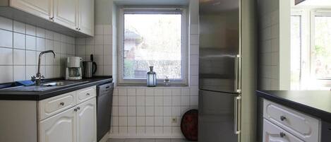 Una cocina pequeña con armarios blancos, un refrigerador de acero inoxidable, una ventana con persianas y piso de baldosas.