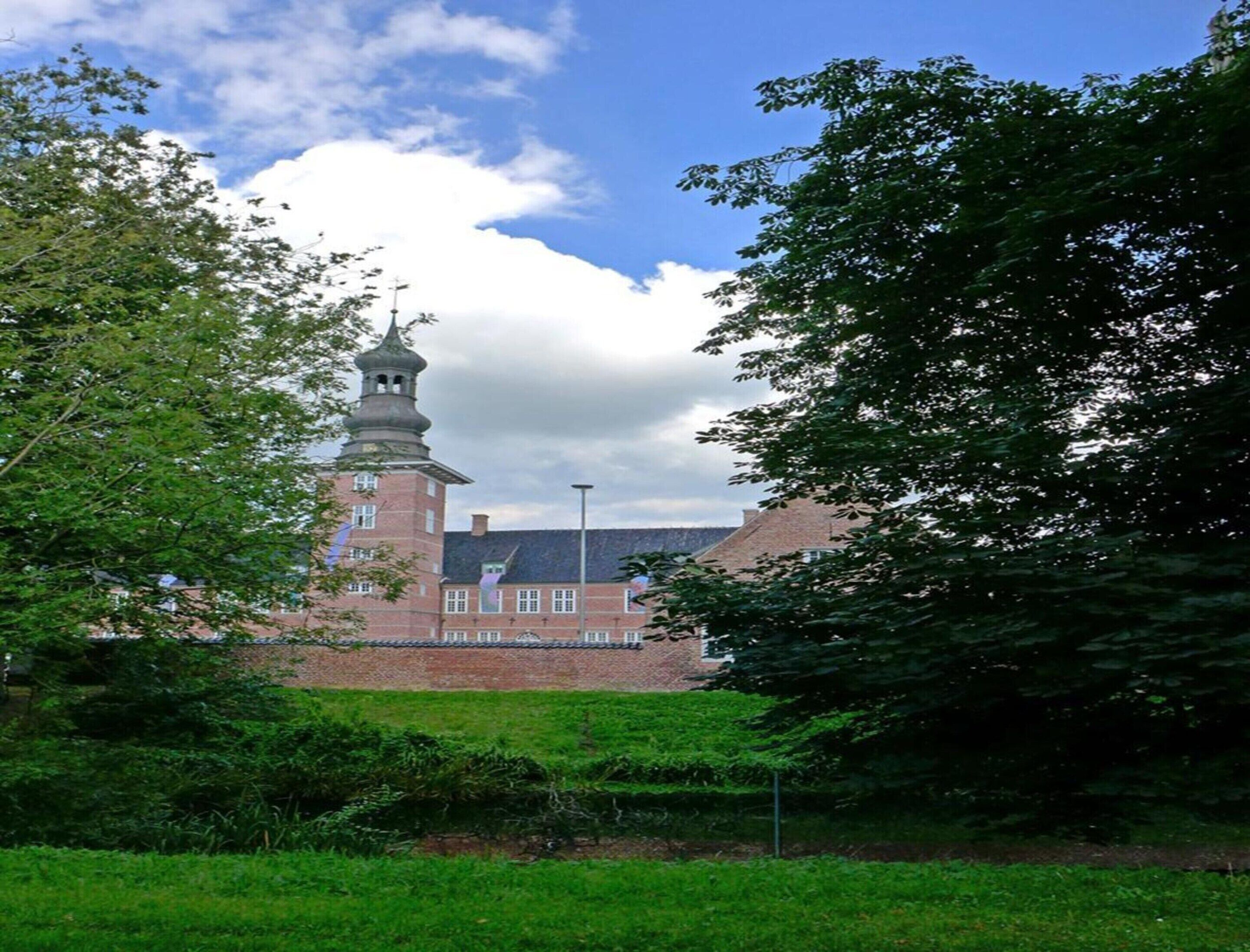 Ein historisches Gebäude mit einem markanten Turm und einer Ziegel-Fassade, umgeben von üppigem Grün.