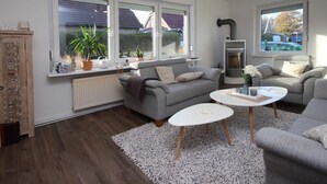 Un salon moderne avec deux canapés gris, une table basse blanche et une cheminée. Des plantes en pot sont posées sur le rebord de la fenêtre, et un meuble décoratif se trouve à gauche.
