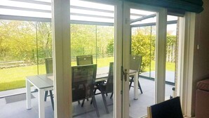 A covered patio with glass doors, outdoor seating, and a view of a green lawn and trees. - Bungalow in Breskens near Beach & Lighthouse (Breskens)