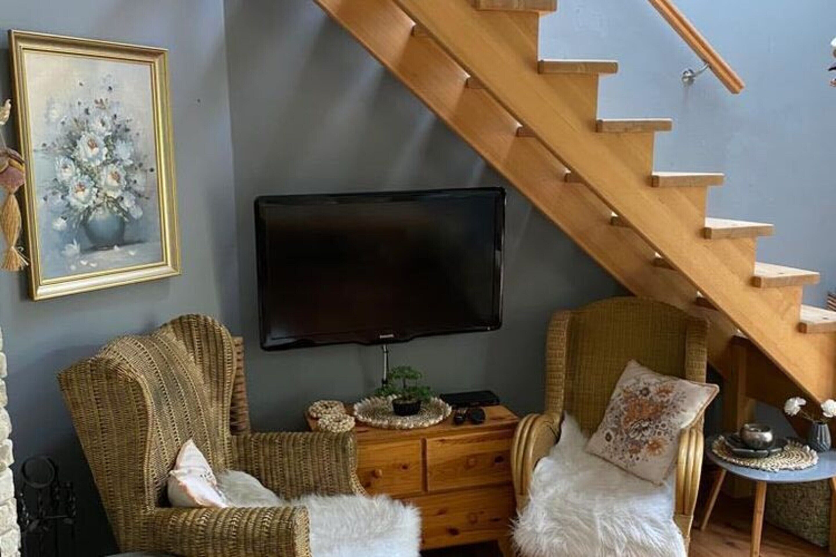 A living room with a wooden staircase, a flat-screen TV, wicker furniture, and a painting on the wall.
