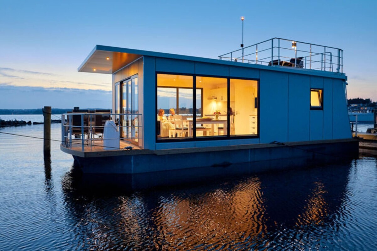 A modern houseboat with a glass exterior, a deck area, and a kitchen visible inside.